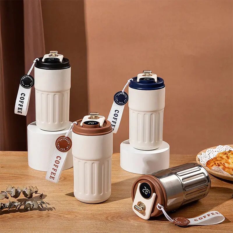 Smart coffee tumblers in white and metallic silver finishes, showing real-time temperature readings on the cap, styled on white display pedestals.